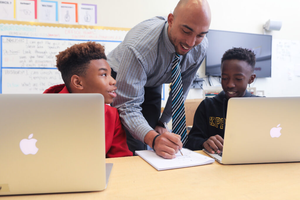 Brandon Nicholson leans over a desk writing on a notebook with two students with Macbooks.