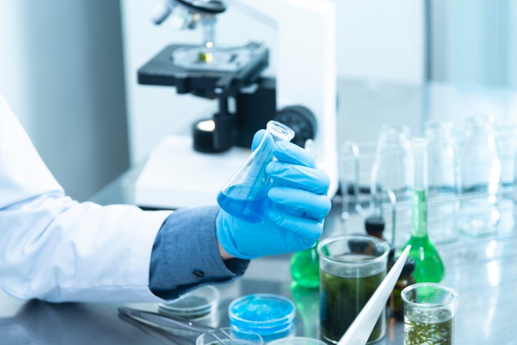 person in lab coat holds flask with microscope nearby