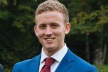 Photo of Dan Bernstein wearing a blue suit jacket, white collared shirt, and red tie. Dan has short cropped curly blonde hair and is smiling while looking at the camera.
